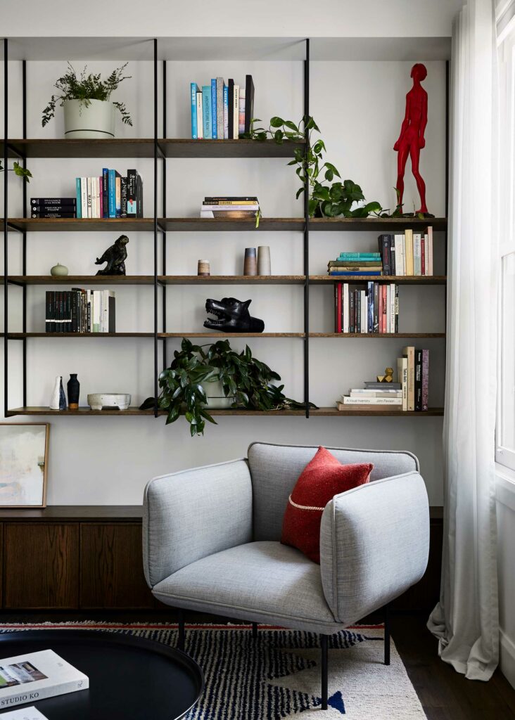 Maike Design lounge. Open bookshelves with black steel frame, decor and plants. Grey armchair and textured woolen rug.